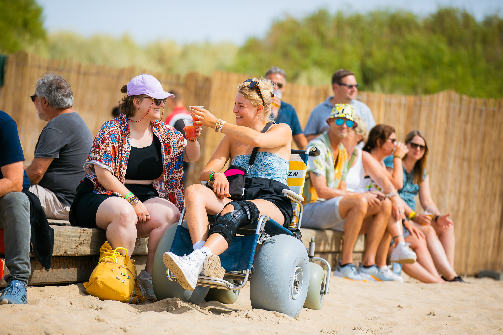 Twee vrouwen proosten op het strand, één van de twee vrouwen zit in een strandrolstoel van HandicapNL