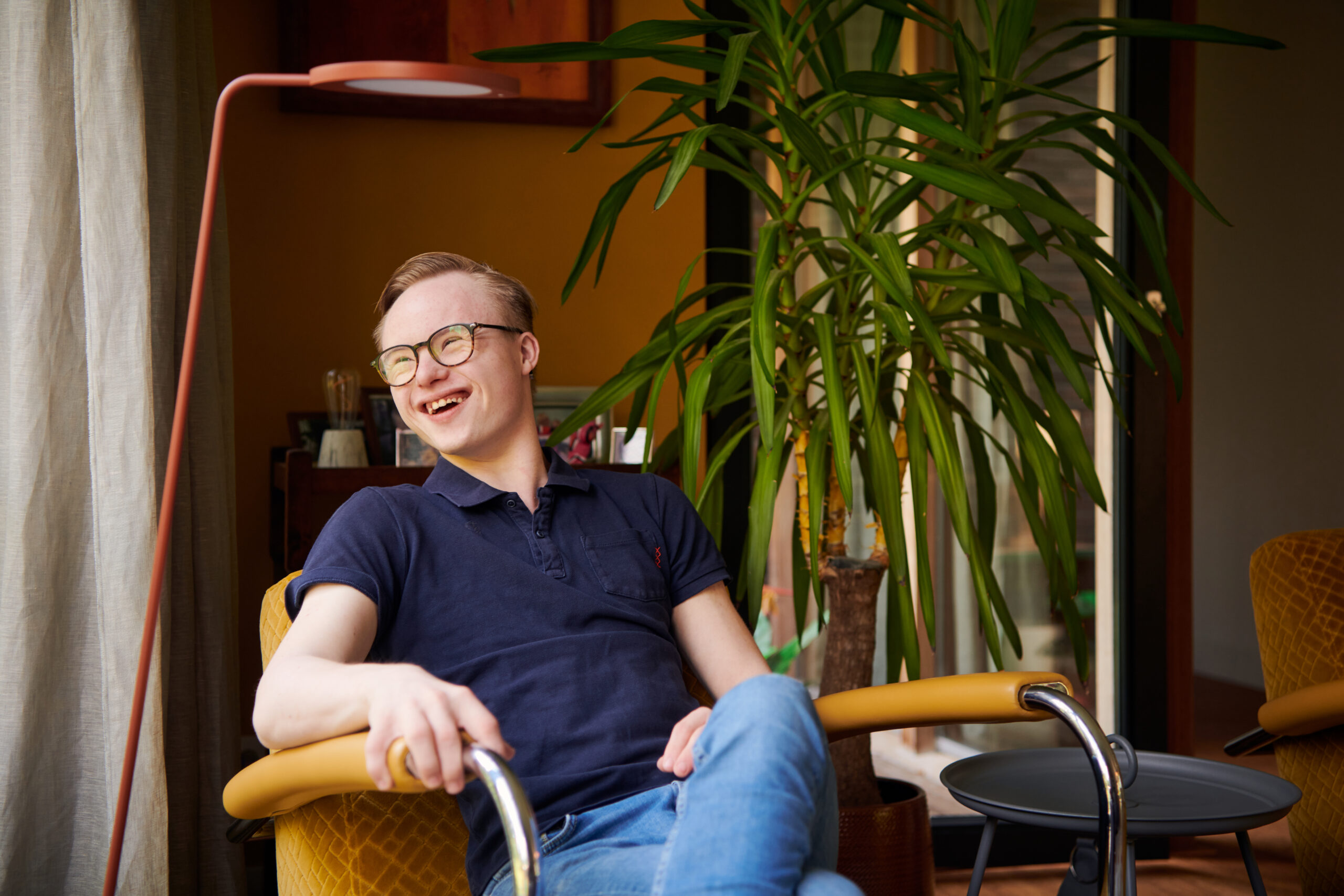 Gijs, een jongen met het Syndroom van Down, zit lachend in een gele fauteuil zit en kijkt naar buiten