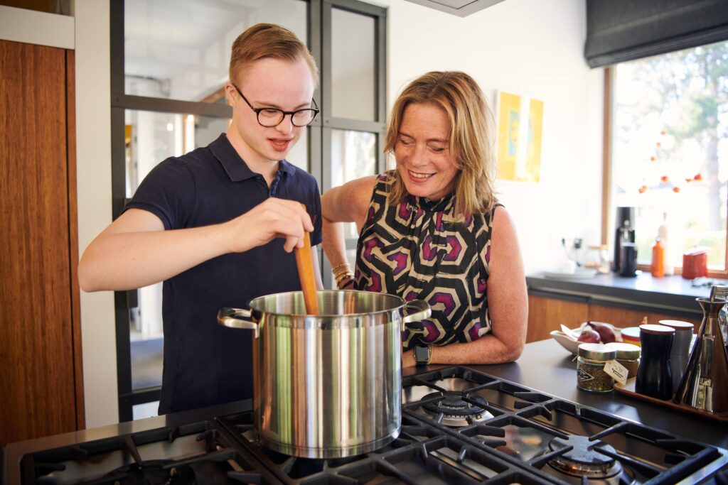 Gijs, een jongen met het Syndroom van Down, staat samen met een vrouw te koken in een keuken. Gijs roert in een grote pan en de vrouw kijkt met hem mee
