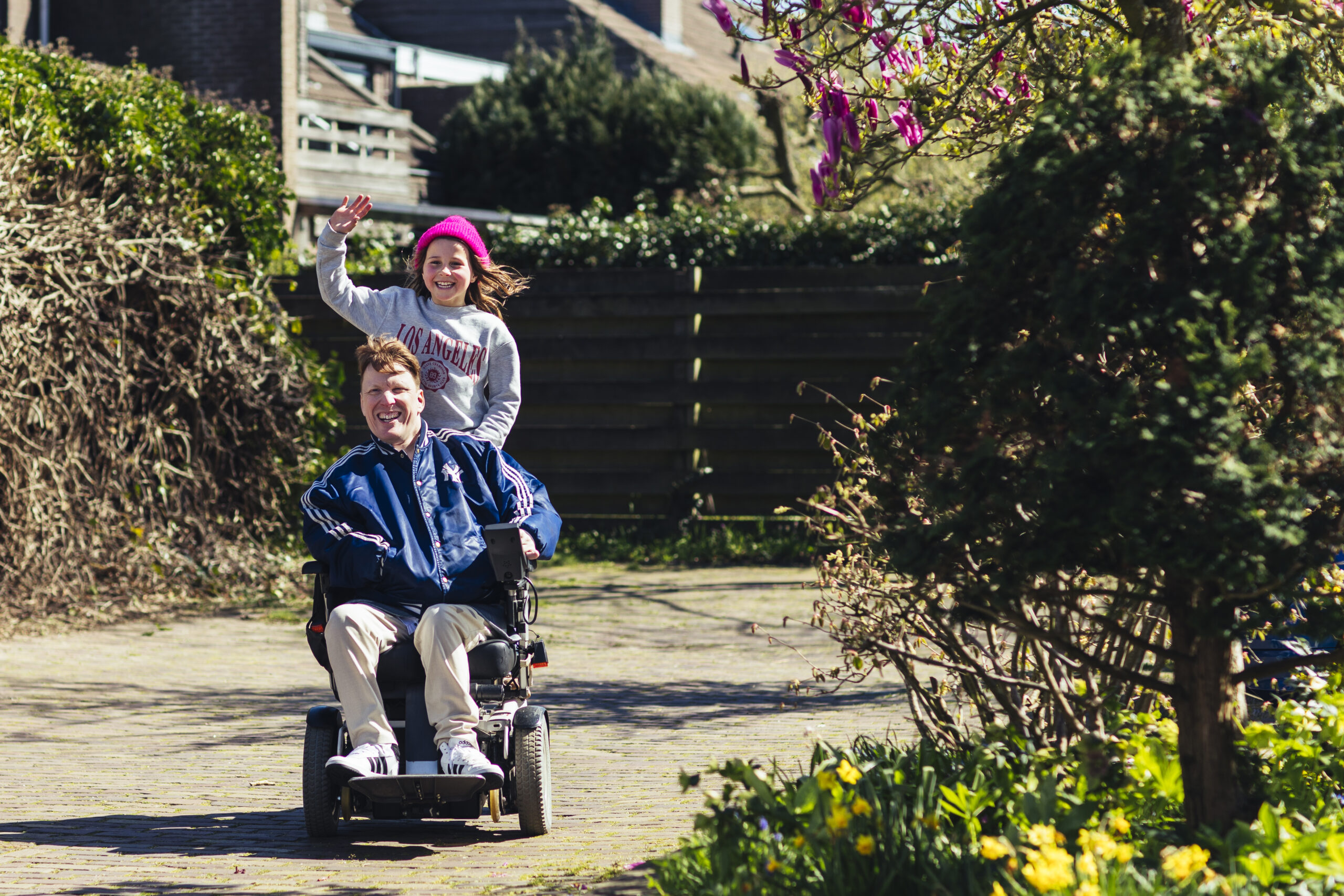 Een meisje zit achterop bij een man die in een rolstoel zit, ze lachen