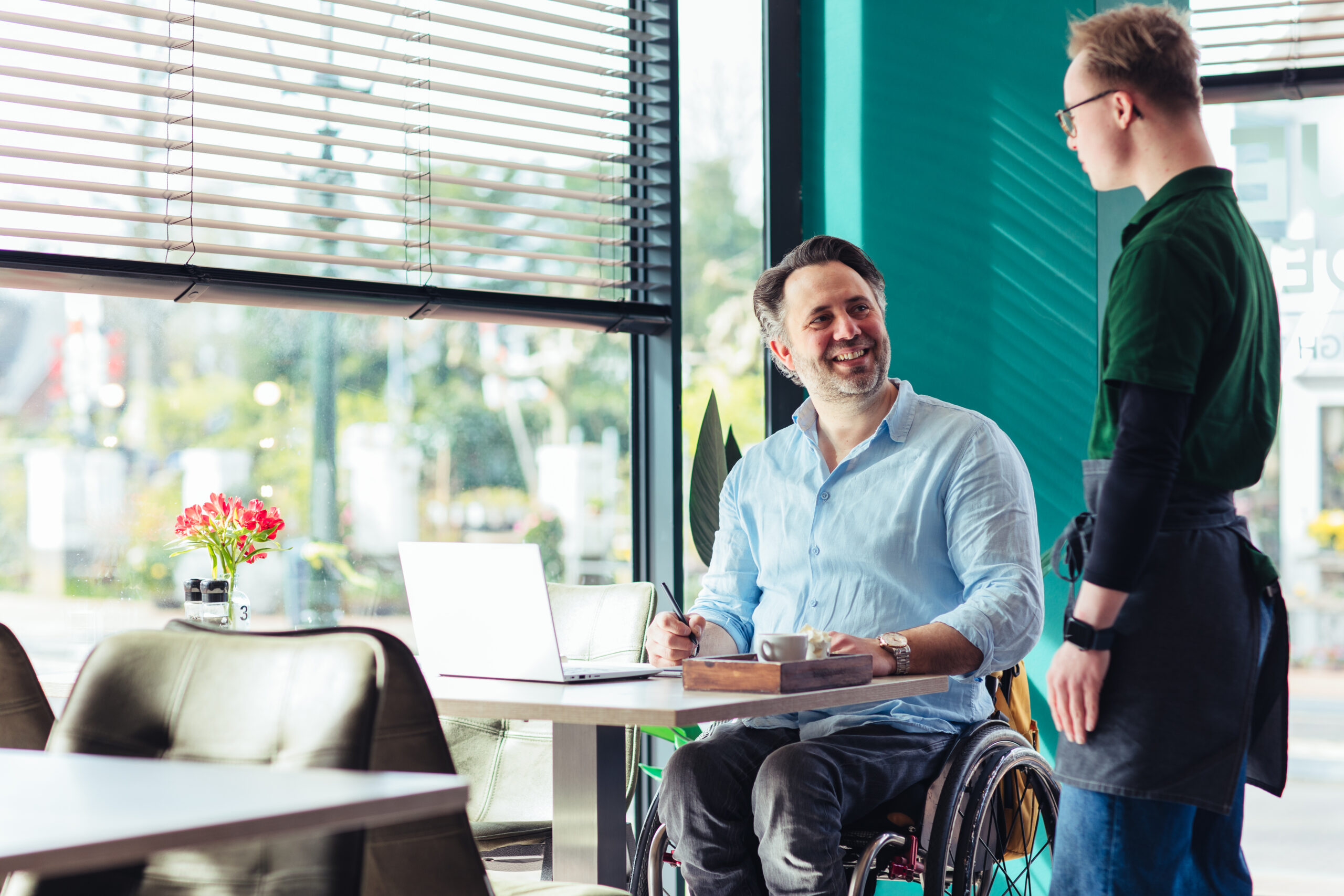 Thomas, een man met een fysieke beperking, zit in zijn rolstoel aan een tafel met zijn laptop en besteld wat bij een medewerker van Lunchroom Beetje Bijzonder