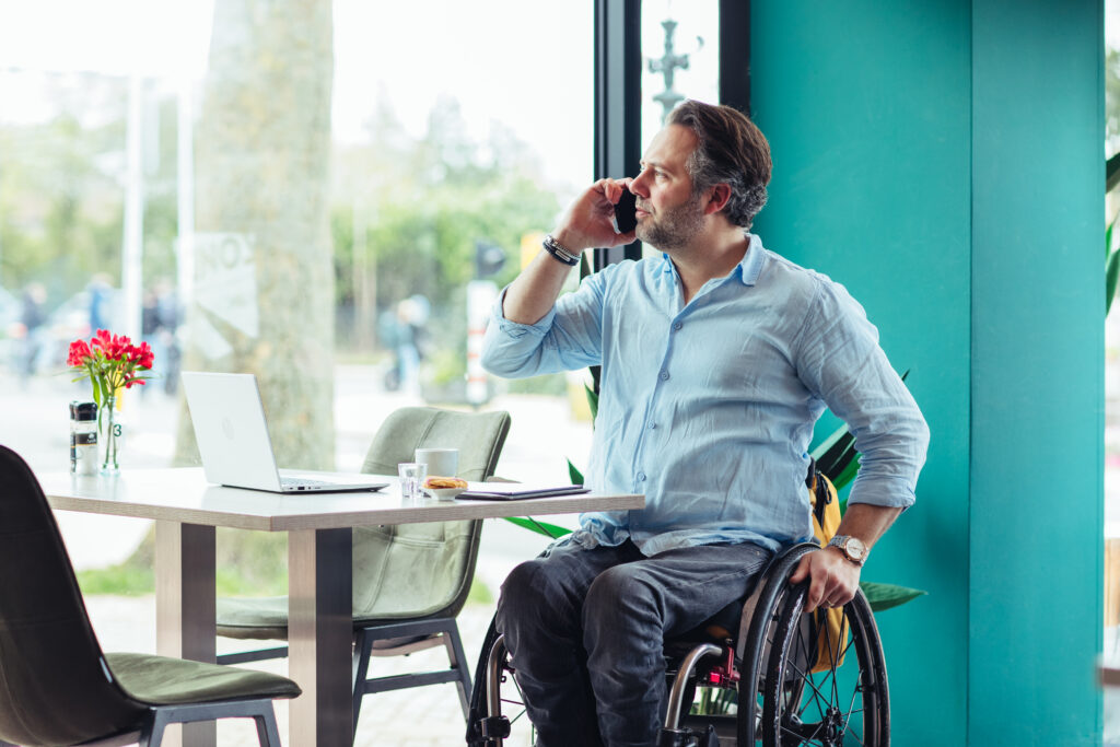 Thomas, een man met een fysieke beperking, zit in zijn rolstoel aan een tafel en pleegt een telefoontje