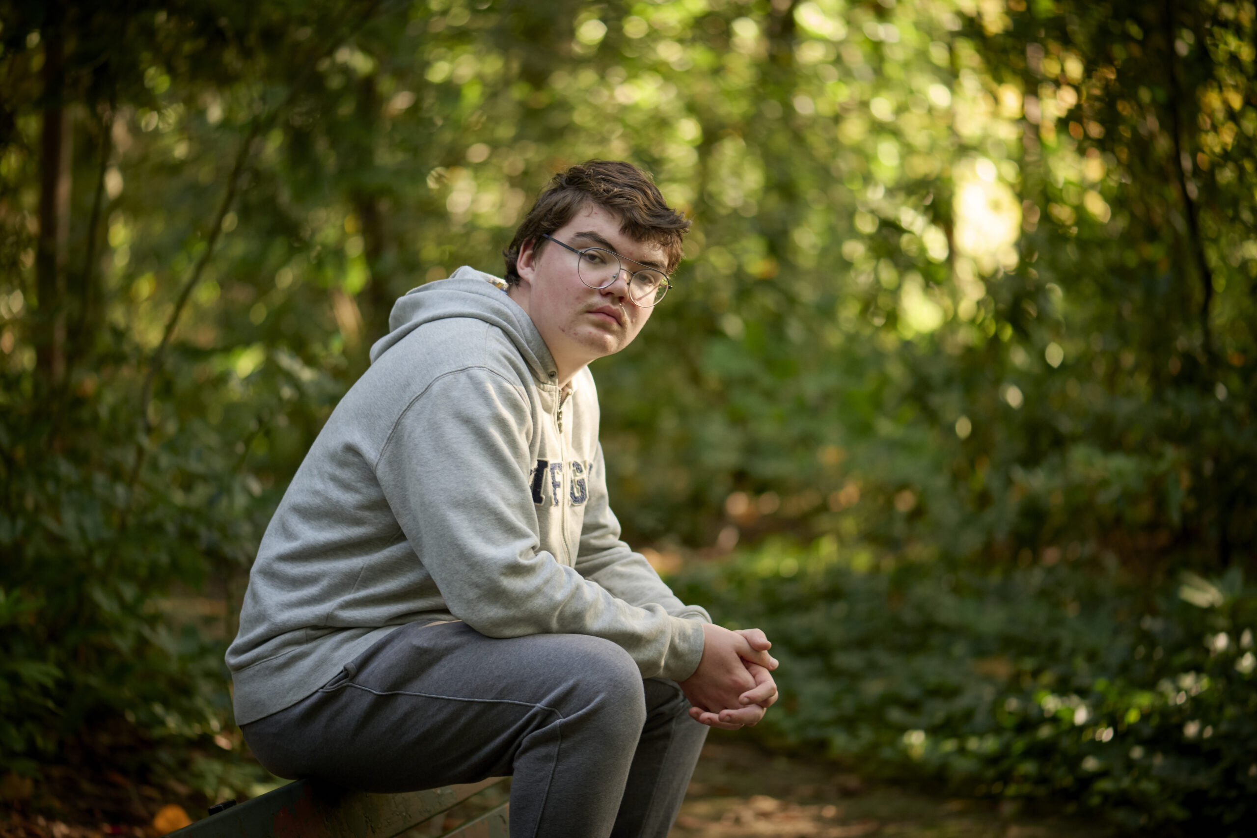 Een jongen met een verstandelijke beperking zit op de rugleuning van een bankje en kijkt je recht aan. Hij zit in een bos, het weer is herfstig.