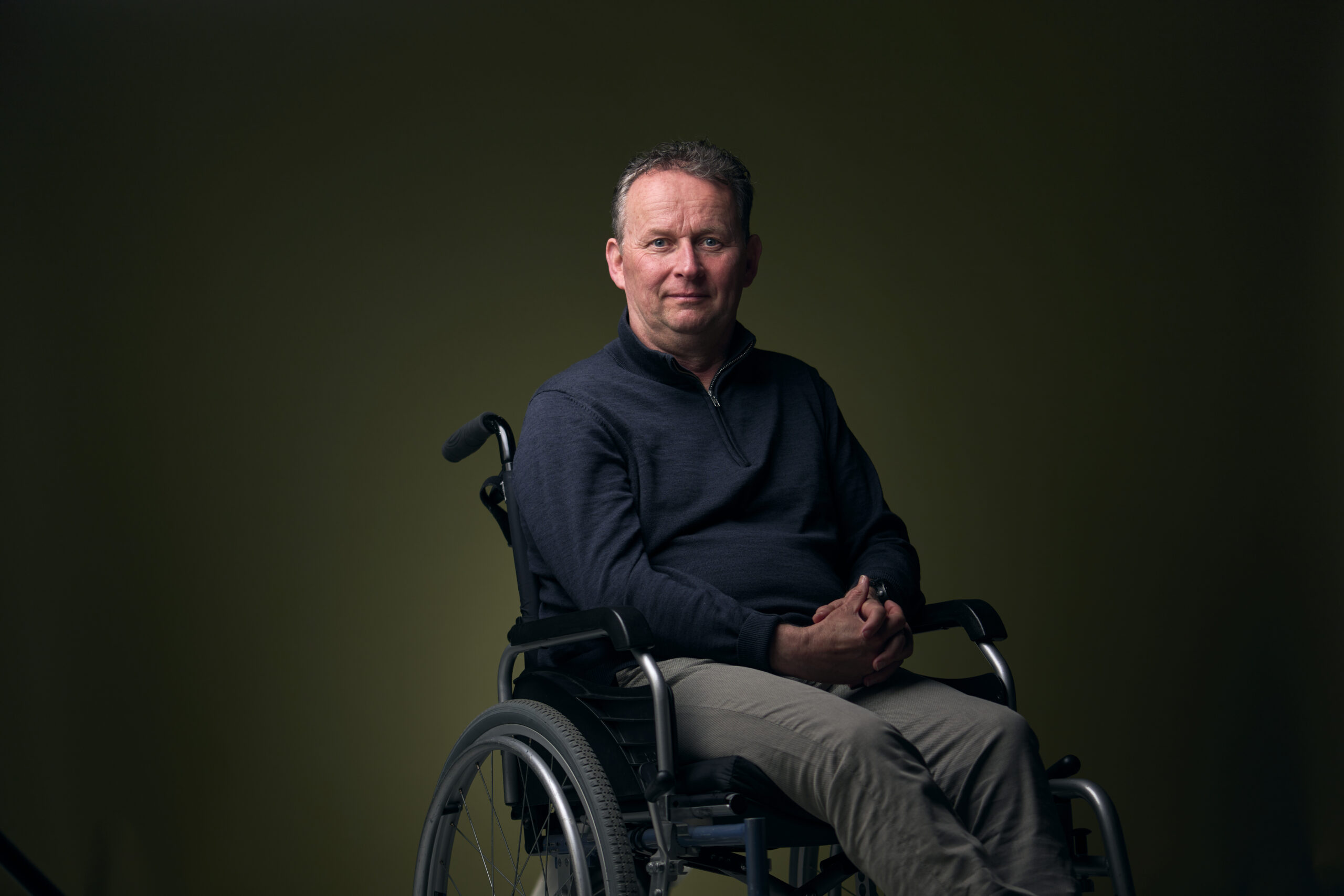 Wim, een man met een fysieke beperking, die in een rolstoel zit en je aankijkt, zijn handen zijn in elkaar gevouwen