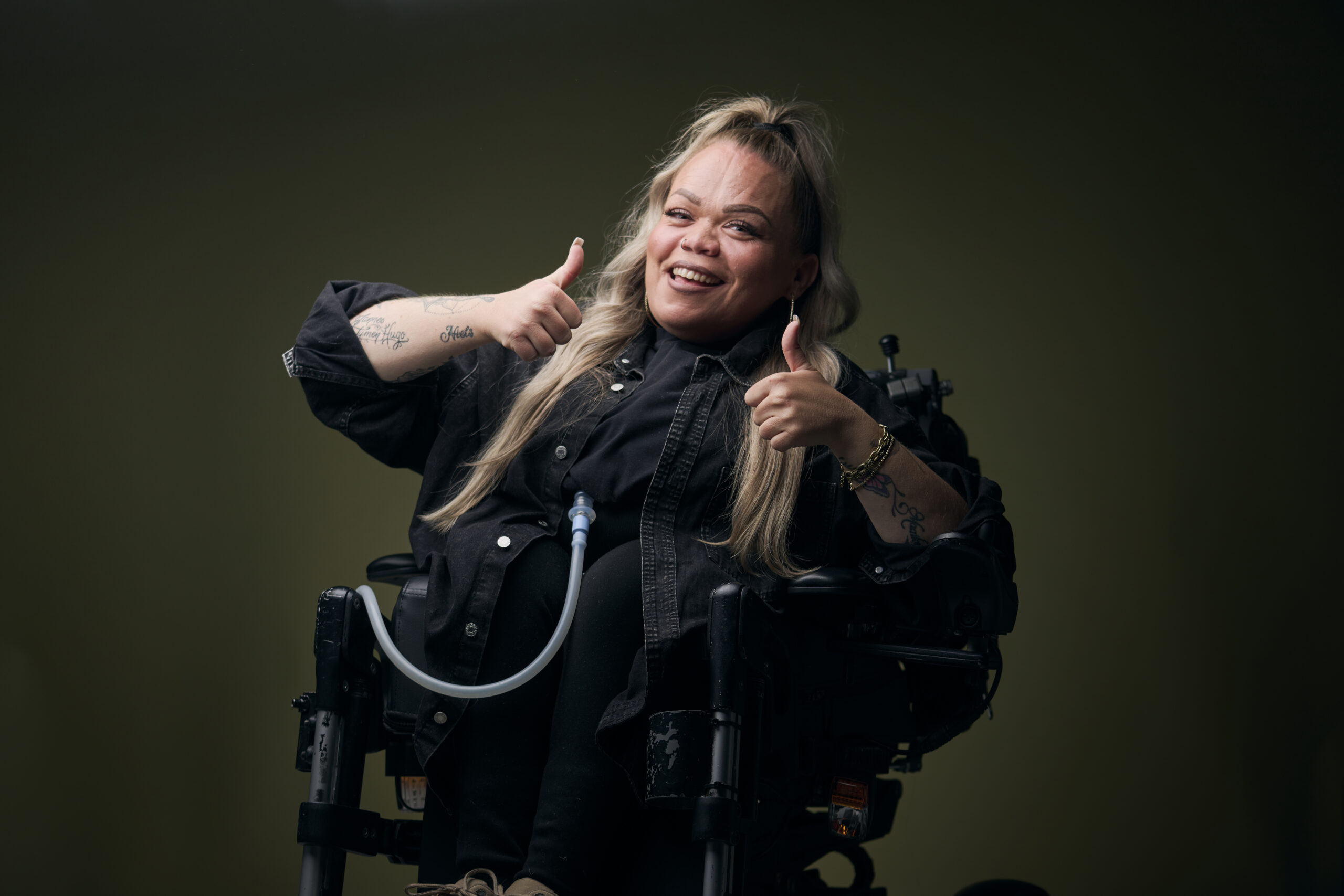 Roos, een vrouw met een fysieke beperking, die in een rolstoel zit en naar je lacht, ze houdt haar duimen omhoog