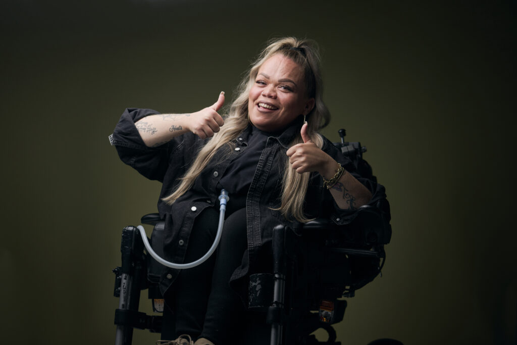 Roos, een vrouw met een fysieke beperking, die in een rolstoel zit en naar je lacht, ze houdt haar duimen omhoog