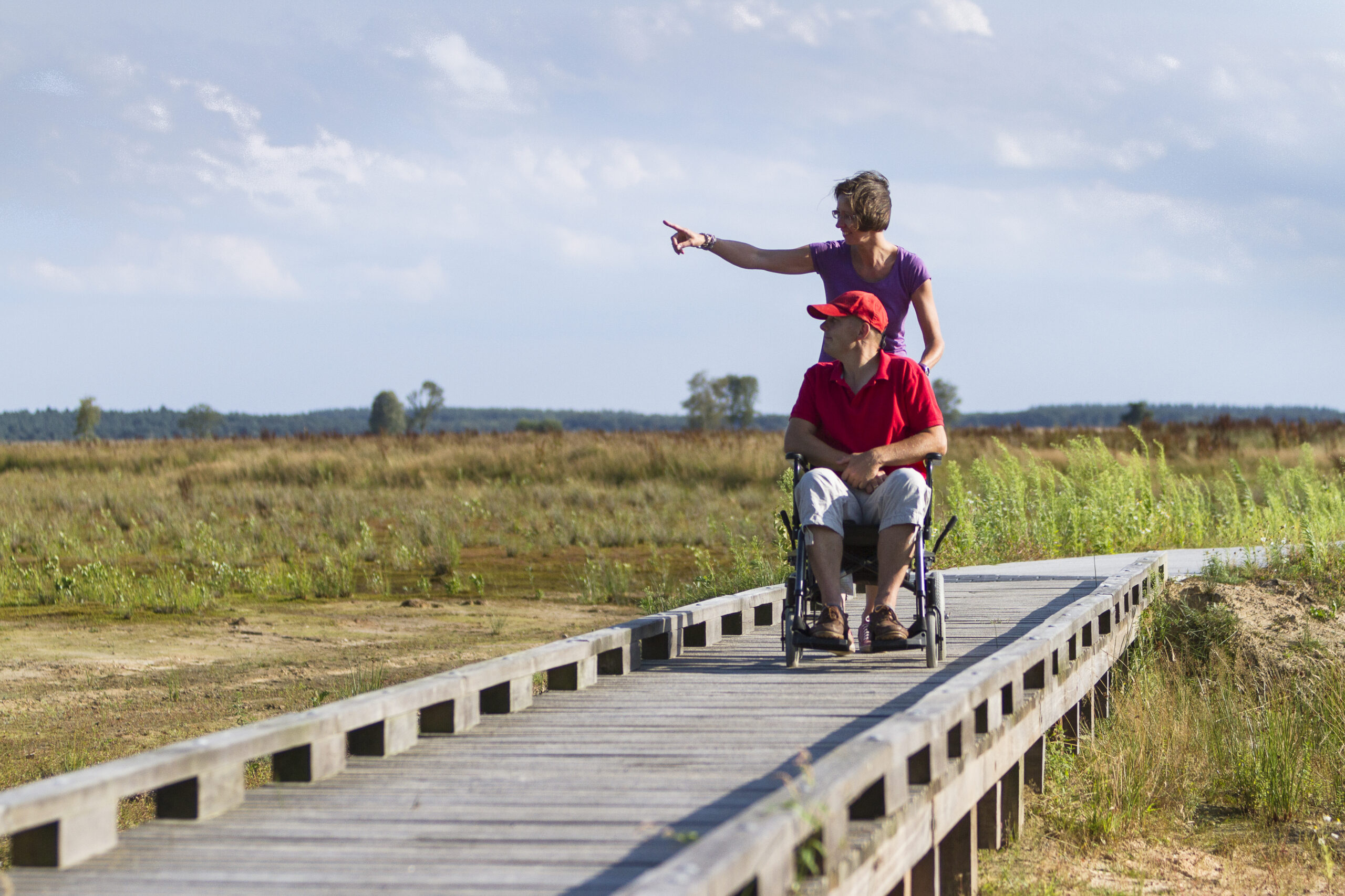Een man in een rolstoel bevind zich samen met een vrouw op een brug middenin een natuurgebied