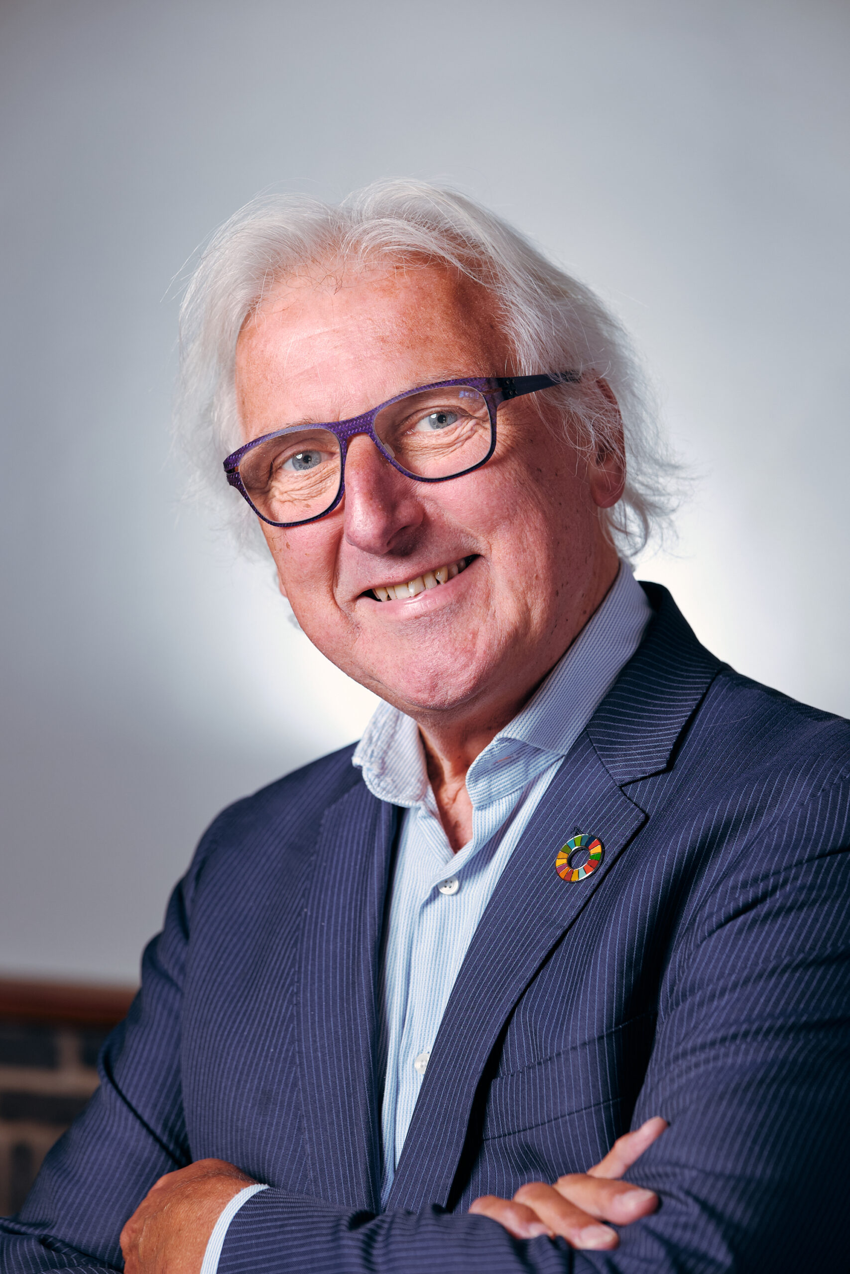 Portret van een oudere man met wit haar en een vriendelijke glimlach. Hij draagt een donkerblauw colbert en een licht overhemd, en heeft een bril met een opvallend paars montuur. Op zijn revers zit een kleurrijke speld. De man kijkt recht in de camera met een open, zelfverzekerde uitstraling, tegen een rustige, lichte achtergrond.