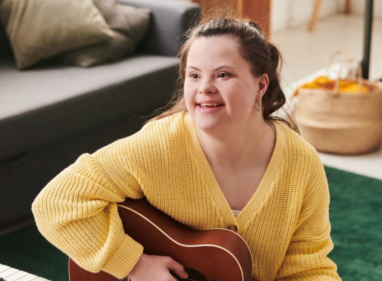Een jong persoon met een verstandelijke beperking zit in een gezellige woonkamer en speelt gitaar. Diegene draagt een gele trui en kijkt met een glimlach opzij. Op de tafel staat een keyboard. De sfeer is warm en ontspannen, met aandacht voor muziek en plezier.