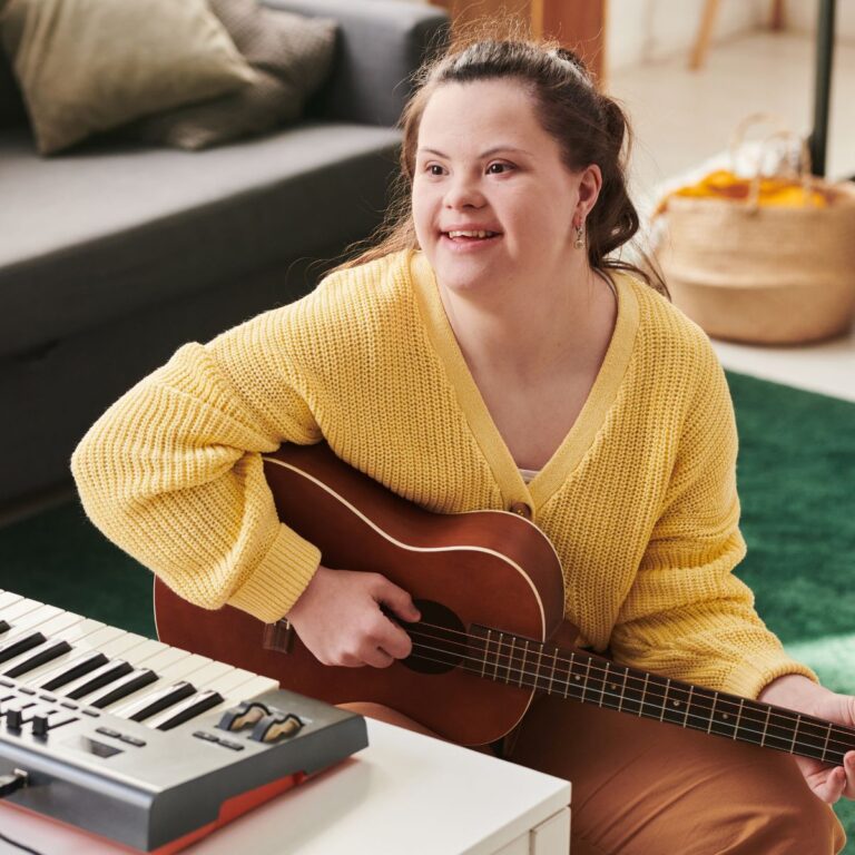 Een jong persoon met een verstandelijke beperking zit in een gezellige woonkamer en speelt gitaar. Diegene draagt een gele trui en kijkt met een glimlach opzij. Op de tafel staat een keyboard. De sfeer is warm en ontspannen, met aandacht voor muziek en plezier.