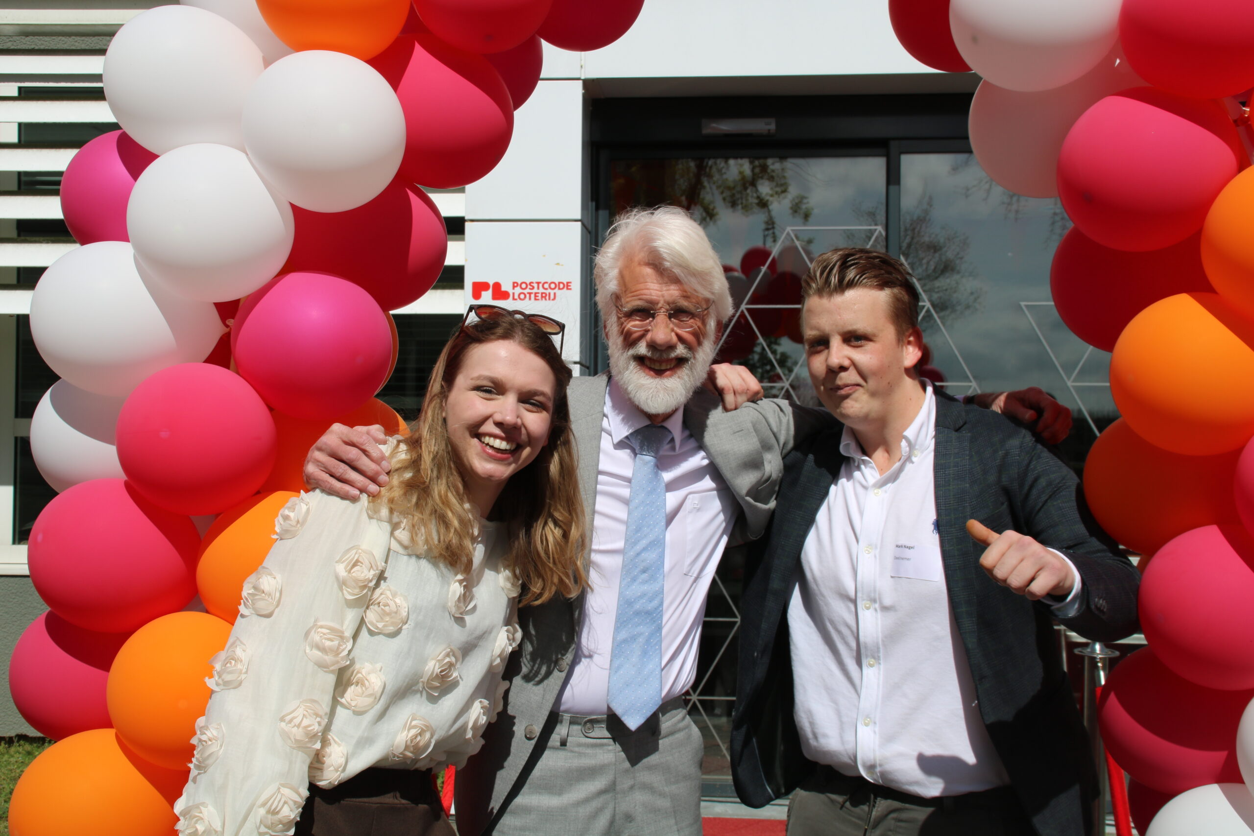 Drie mensen staan lachend samen onder een boog van roze, witte en oranje ballonnen bij de ingang van een gebouw. In het midden staat een oudere man met wit haar en baard, met zijn armen om twee jonge volwassenen heen. Op de achtergrond is een ingang te zien met een logo van de Postcode Loterij.