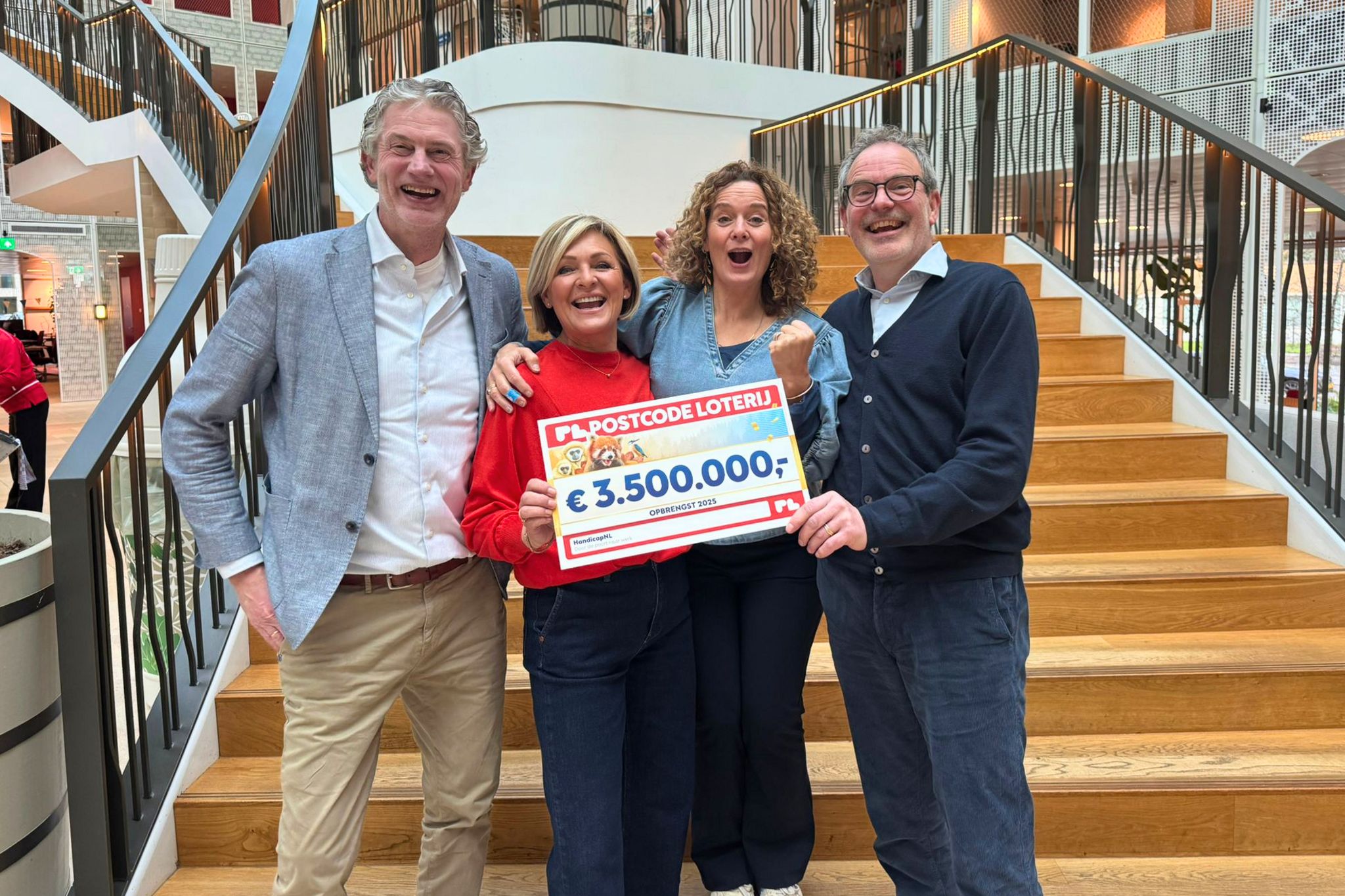 Vier mensen staan samen op een brede trap in een modern gebouw en lachen enthousiast naar de camera. Ze houden samen een grote cheque vast met een bedrag van €3.500.000, wat wijst op een feestelijk moment of donatie. De groep staat dicht bij elkaar met armen om elkaar heen, wat verbondenheid en blijdschap uitstraalt.