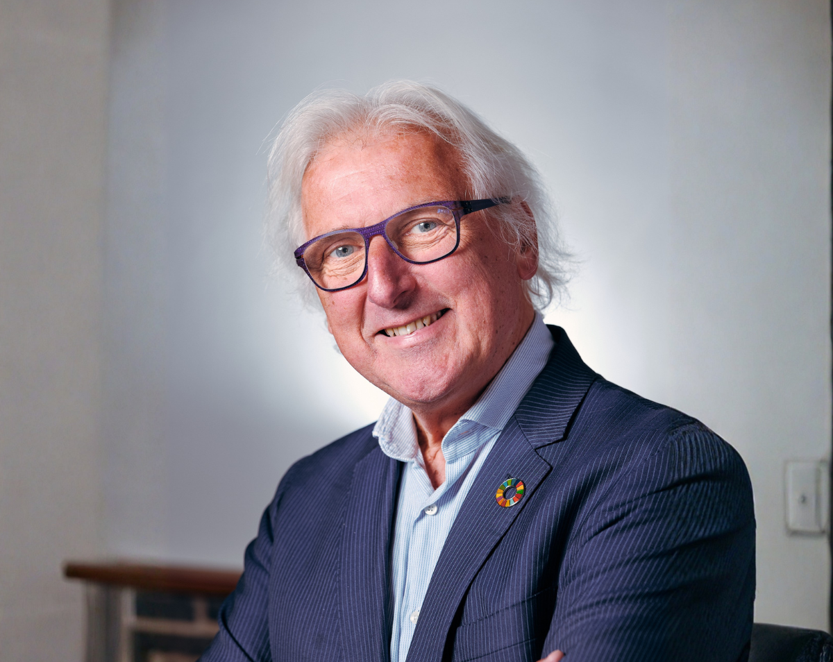 Portret van een oudere man met wit haar en een vriendelijke glimlach. Hij draagt een donkerblauw colbert en een licht overhemd, en heeft een bril met een opvallend paars montuur. Op zijn revers zit een kleurrijke speld. De man kijkt recht in de camera met een open, zelfverzekerde uitstraling, tegen een rustige, lichte achtergrond.