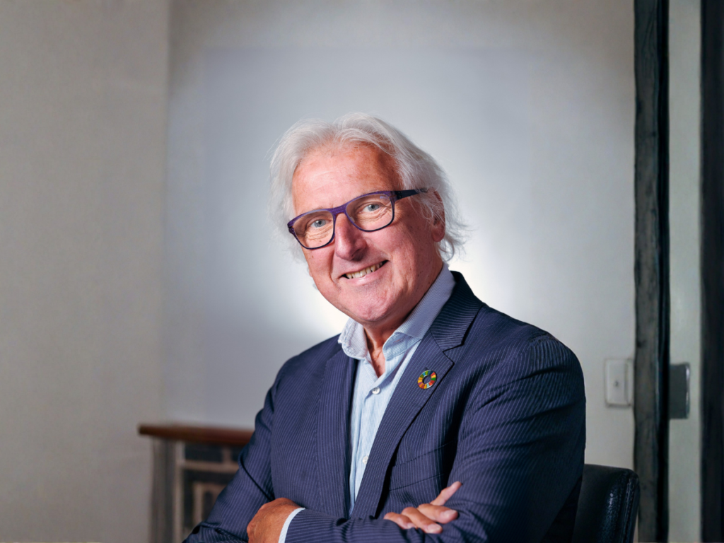 Portret van een oudere man met wit haar en een vriendelijke glimlach. Hij draagt een donkerblauw colbert en een licht overhemd, en heeft een bril met een opvallend paars montuur. Op zijn revers zit een kleurrijke speld. De man kijkt recht in de camera met een open, zelfverzekerde uitstraling, tegen een rustige, lichte achtergrond.