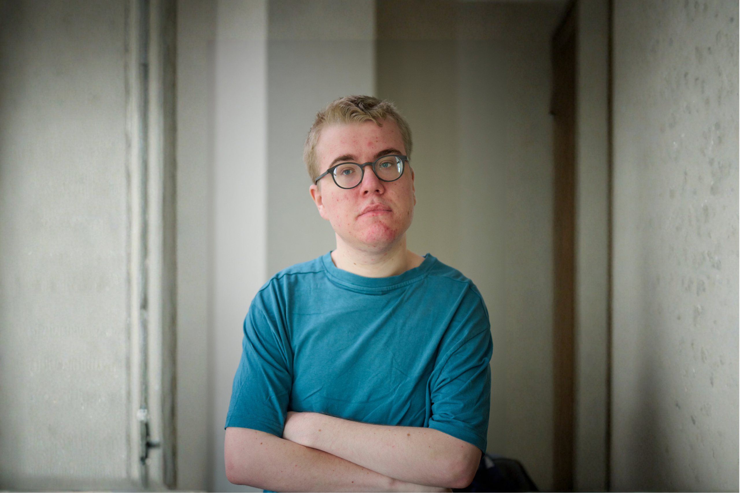 Jongere met een lichte huidskleur en kort blond haar staat binnen tegen een muur, met de armen over elkaar. Diegene draagt een turquoise T-shirt en een bril met ronde glazen en kijkt recht in de camera met een neutrale, serieuze blik.