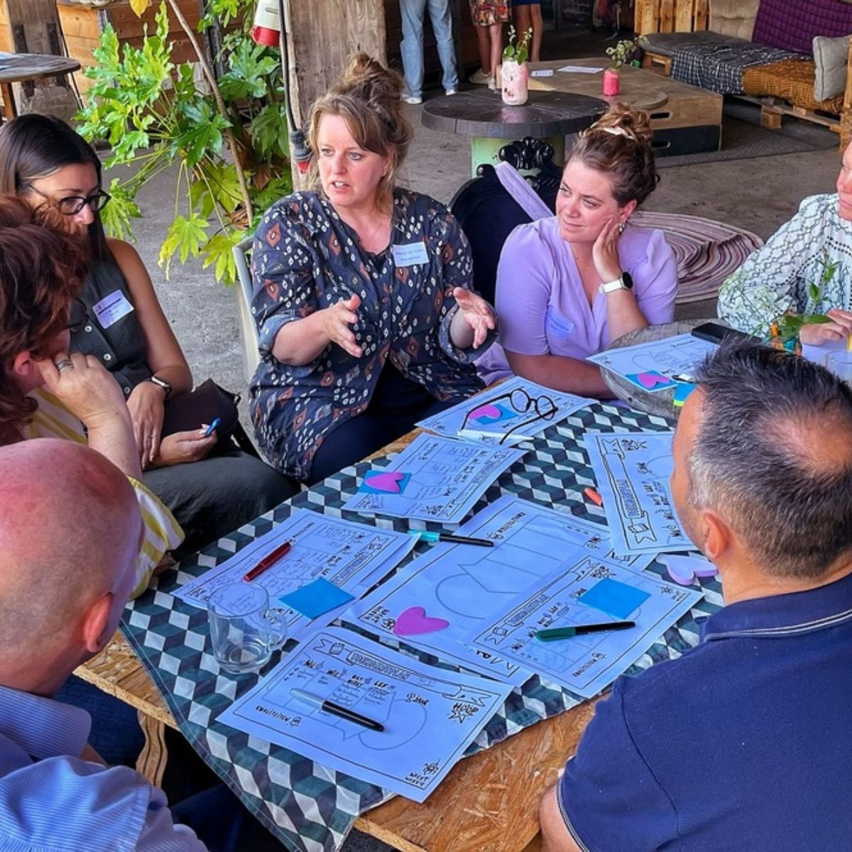 Deelnemers aan een werksessie zitten rond een tafel en gaan met elkaar in gesprek. Op tafel liggen werkbladen, stiften en post-its voor een gezamenlijke brainstorm.