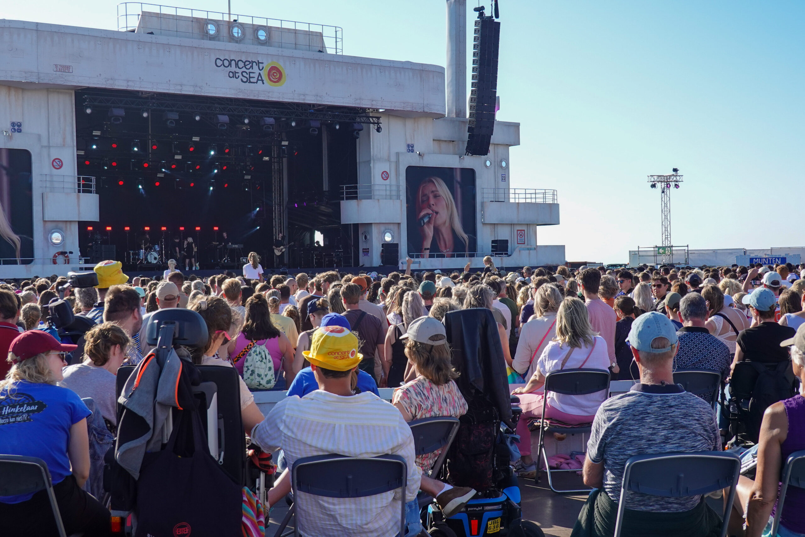 Het podium van Concert at Sea is zichtbaar vanaf het verhoogde podium, mensen kijken naar het optreden dat plaatsvindt