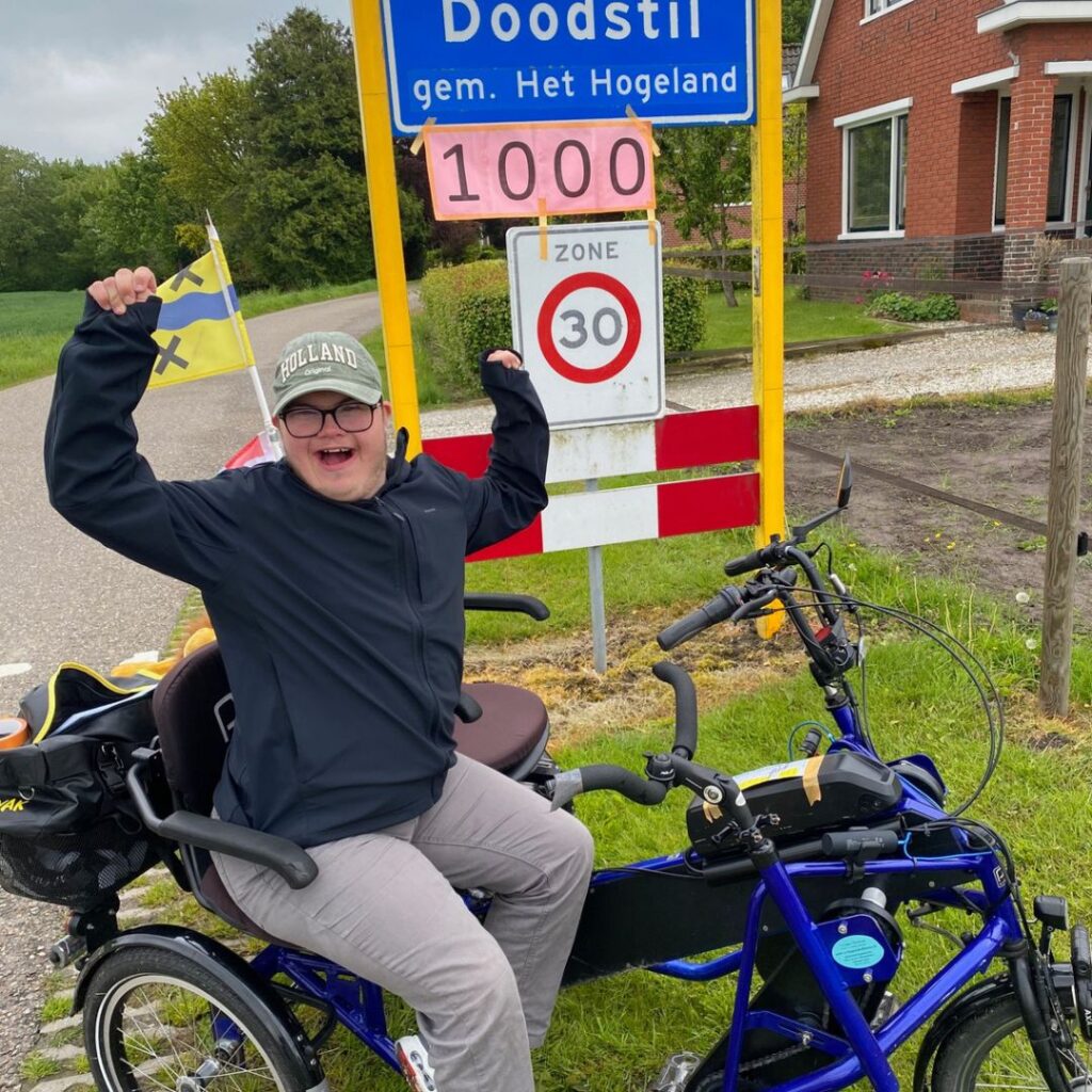 Juichende persoon op een aangepaste driewielfiets bij het plaatsnaambord van Doodstil, met een roze bordje waarop ‘1000’ staat. Op de achtergrond een landelijke omgeving met een huis en bomen.