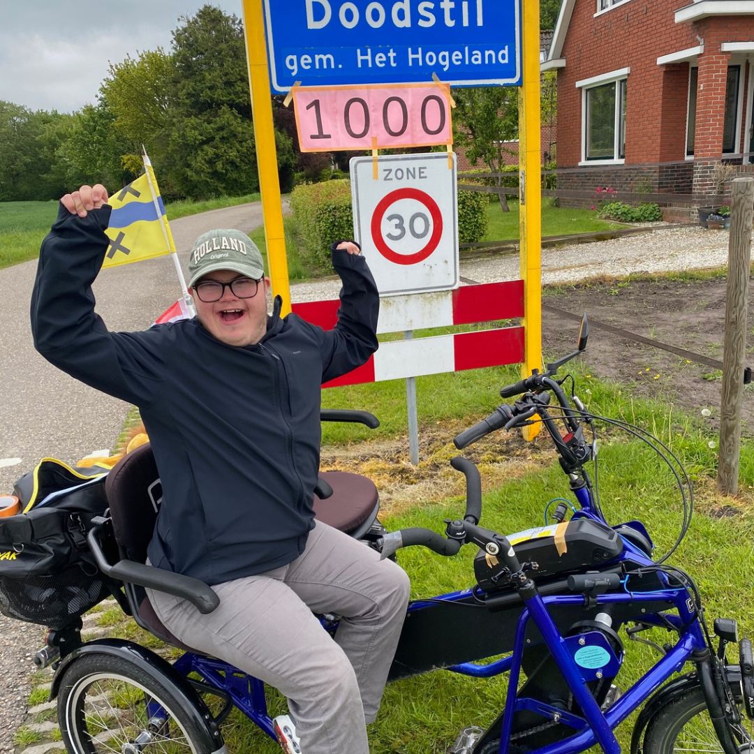 Juichende persoon op een aangepaste driewielfiets bij het plaatsnaambord van Doodstil, met een roze bordje waarop ‘1000’ staat. Op de achtergrond een landelijke omgeving met een huis en bomen.
