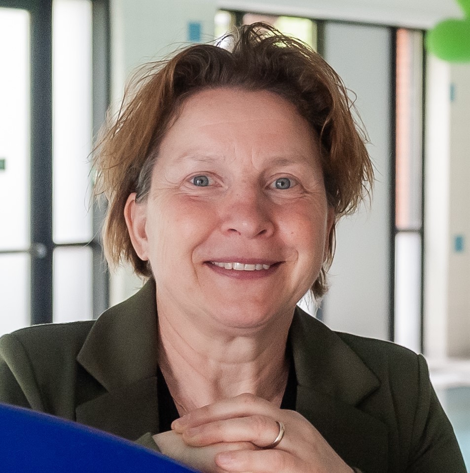 Portret van Karin den Balvert in een lichte binnenruimte. Karin kijkt recht in de camera en glimlacht. Op de achtergrond zijn ramen en een onscherpe kantoor- of ontmoetingsruimte zichtbaar.