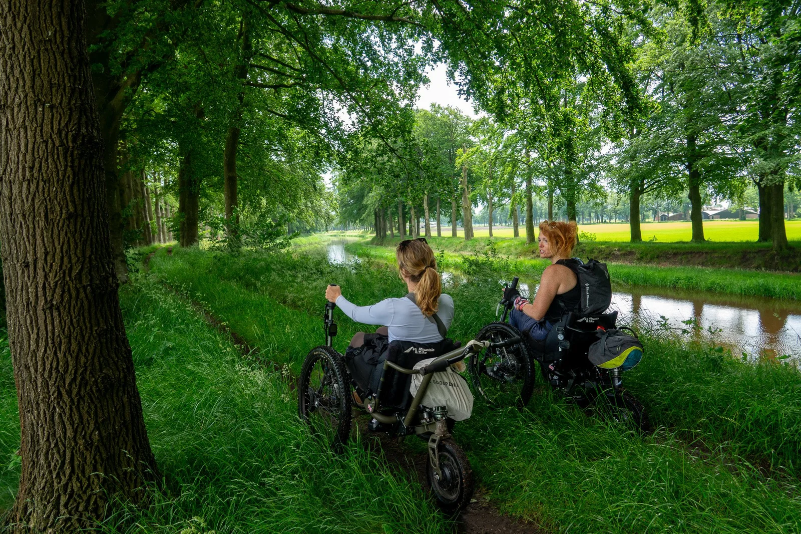 Twee vrouwen rijden in aangepaste terreinrolstoelen over een smal bospad langs een kanaal. Ze genieten van de groene omgeving en kijken uit over het landschap.