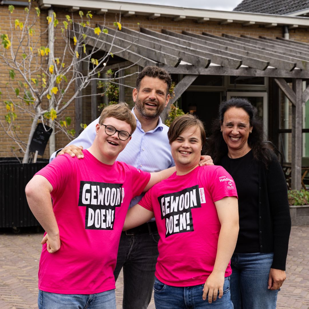 Vier mensen staan lachend naast elkaar voor de ingang van het Lotus College in Haarlem. Twee jongvolwassenen dragen een felroze T-shirt met de tekst 'Gewoon Doen!'. Achter hen staan een vrouw en een man met hun armen om de jongeren heen