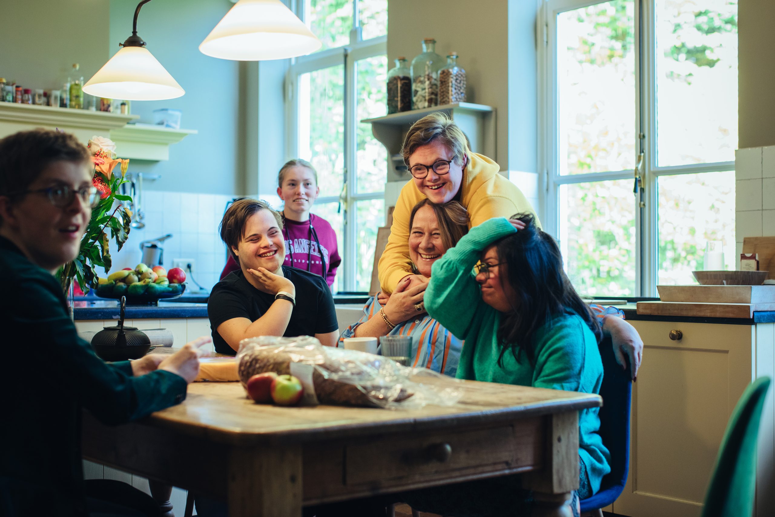 Een groep jongeren en volwassenen zit samen rond een keukentafel. Ze lachen en maken plezier met elkaar in een lichte, huiselijke keuken.
