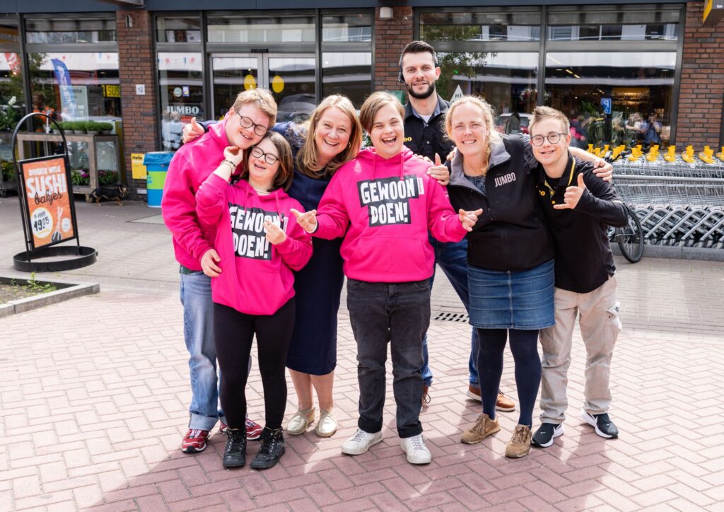 Een groep van acht mensen poseert lachend voor de Jumbo in Driebergen. Drie van hen dragen felroze truien met de tekst 'Gewoon Doen!'. De anderen dragen vrijetijdskleding of Jumbo-werkkleding. Ze staan dicht bij elkaar en maken vrolijke handgebaren.