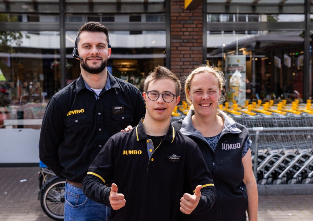 Jan staat lachend tussen twee collega's voor de Jumbo in Driebergen. Hij draagt een zwart Jumbo-poloshirt, een bril en steekt beide duimen omhoog. Zijn collega's staan aan weerszijden van hem en dragen ook Jumbo-werkkleding.