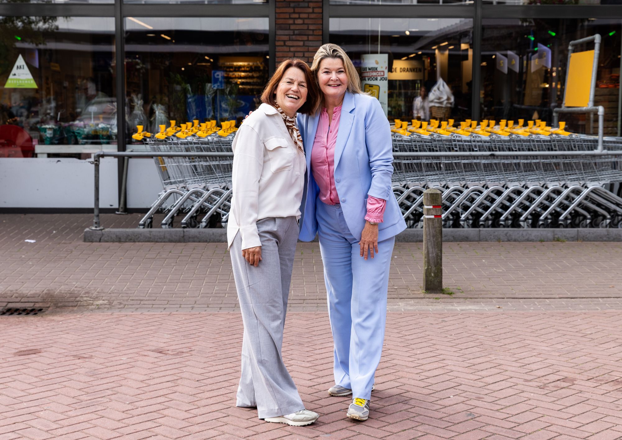Twee lachende vrouwen staan met hun armen om elkaar heen voor de ingang van Jumbo in Driebergen. De ene vrouw draagt een witte blouse en een lichtgrijze broek, de andere een lichtblauw pak met een roze blouse.